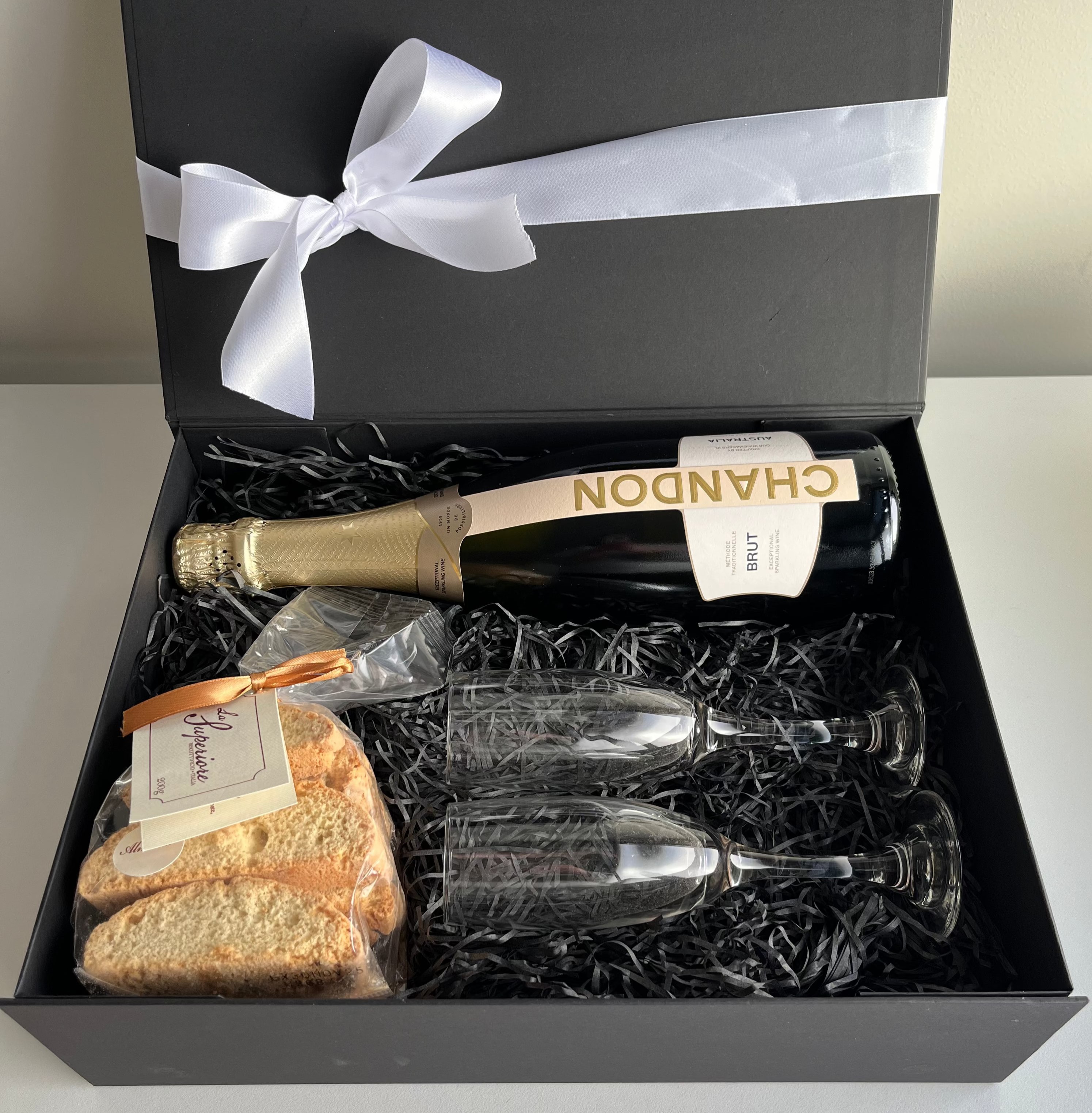Gift set with champagne, flutes, and cantucci in a black box with a white ribbon.